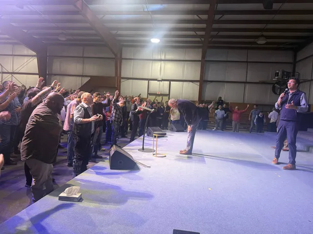 Men pray at the stage while a platform leader addresses the room.